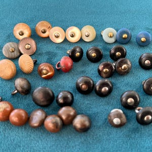 May include: A selection of buttons in diverse colours and designs. The buttons feature shades of brown, black, blue, and beige. Some have a metallic finish, while others have a smooth, matte appearance. The buttons are arranged on a teal fabric.