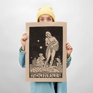 May include: A person is holding up a brown poster with a black and white illustration of a man holding a pitcher of water. The poster has the word "Aquarius" printed at the bottom.