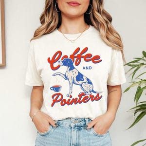 May include: Cream-colored t-shirt with the text "Coffee and Pointers" in red and blue. The graphic features a blue and white dog with a coffee cup. The shirt is a casual, everyday wear item.