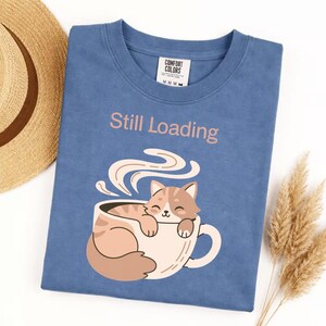 May include: A blue t-shirt with the words "Still Loading" printed in a light brown color. The shirt features a graphic of a cat relaxing in a coffee cup. The cat is light brown and white.