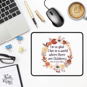 May include: White rectangular mousepad with a black border, featuring a floral wreath design and the quote "I'm so glad I live in a world where there are Octobers" from Anne of Green Gables. A computer mouse, pen, and coffee cup are also visible.