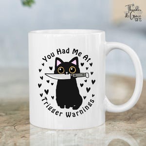 Può includere: Tazza in ceramica bianca con un gatto nero che tiene un coltello. La tazza presenta la scritta "You Had Me At Trigger Warnings" in un design circolare con cuori. La tazza ha un manico bianco e le parole "Thunder & Grace GIFTS" sono stampate a destra.