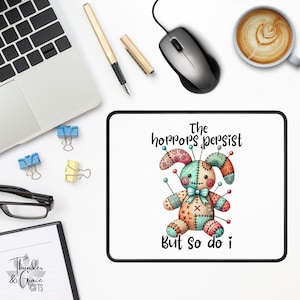 May include: A white mouse mat with a black border, displaying a stitched bunny design with pins and the text "The horrors persist but so do I." A laptop, mouse, pen, coffee cup, and office supplies are also visible.