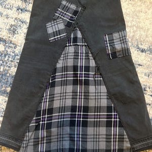May include: A dark gray denim skirt with a plaid fabric panel and plaid patches. The plaid pattern includes shades of gray, black, and purple. The skirt has a unique, patchwork design.