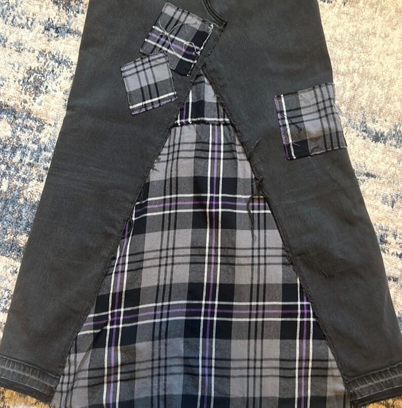 May include: A dark gray denim skirt with a plaid fabric panel and plaid patches. The plaid pattern includes shades of gray, black, and purple. The skirt has a unique, patchwork design.