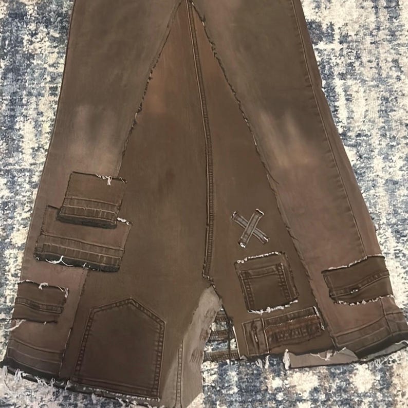 May include: Brown denim skirt with a patchwork design. The skirt features multiple pockets, distressed edges, and an X-shaped detail. The skirt has a unique, asymmetrical design.
