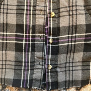 May include: Close-up of a button-down shirt with a plaid pattern in shades of gray, black, white, and purple. The shirt has visible buttons and a frayed hem, suggesting a vintage or distressed style. The fabric appears to be a soft, woven material.