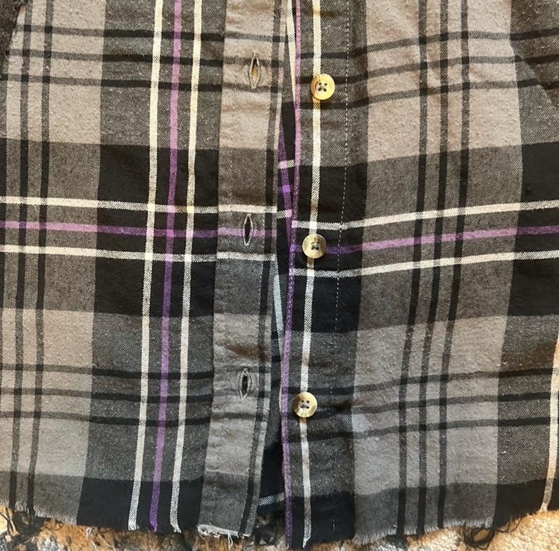 May include: Close-up of a button-down shirt with a plaid pattern in shades of gray, black, white, and purple. The shirt has visible buttons and a frayed hem, suggesting a vintage or distressed style. The fabric appears to be a soft, woven material.