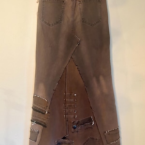 May include: A brown denim skirt with a unique patchwork design. The skirt features a high waist, back pockets, and a split hem. Patches of denim are sewn together, creating a distressed and textured look. The skirt is hung on a wooden hanger.
