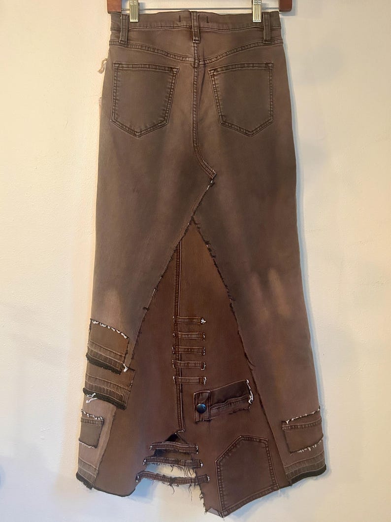 May include: A brown denim skirt with a unique patchwork design. The skirt features a high waist, back pockets, and a split hem. Patches of denim are sewn together, creating a distressed and textured look. The skirt is hung on a wooden hanger.