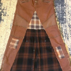 May include: A brown denim skirt with a patchwork design. The skirt features a button closure, side zippers, and plaid fabric inserts. Patches of plaid fabric are sewn onto the denim. The skirt has a unique, repurposed aesthetic.