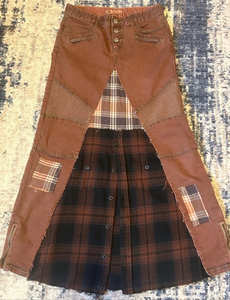 May include: A brown denim skirt with a patchwork design. The skirt features a button closure, side zippers, and plaid fabric inserts. Patches of plaid fabric are sewn onto the denim. The skirt has a unique, repurposed aesthetic.