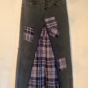 May include: A dark gray denim skirt with a patchwork design. The skirt features a plaid fabric panel in the center and several plaid patches. The skirt has a button closure and zipper details.