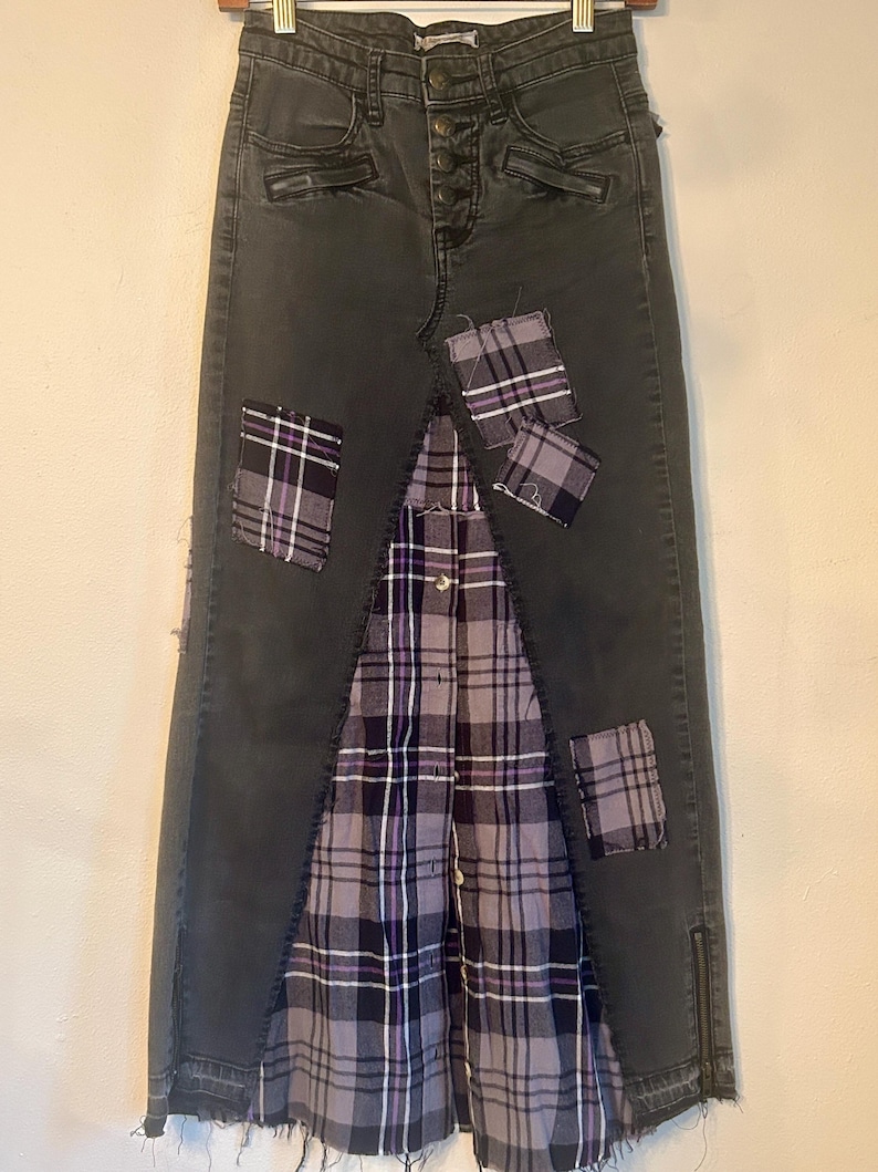 May include: A dark gray denim skirt with a patchwork design. The skirt features a plaid fabric panel in the center and several plaid patches. The skirt has a button closure and zipper details.