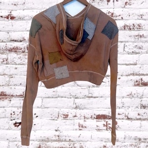 Może przedstawiać: Brązowa krótka bluza z kapturem o strukturze wafelka. Bluza z kapturem posiada naszywki w różnych kolorach i fakturach, w tym szarym, zielonym i niebieskim. Bluza z kapturem ma długie rękawy i kaptur. Odzież wisi na drewnianym wieszaku na tle białej ceglanej ściany.