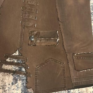 May include: Close-up of a brown jacket with multiple pockets and decorative stitching. The jacket has a distressed look with frayed edges and a button closure on one of the pockets. The fabric appears to be a sturdy material, possibly denim or a similar textile.