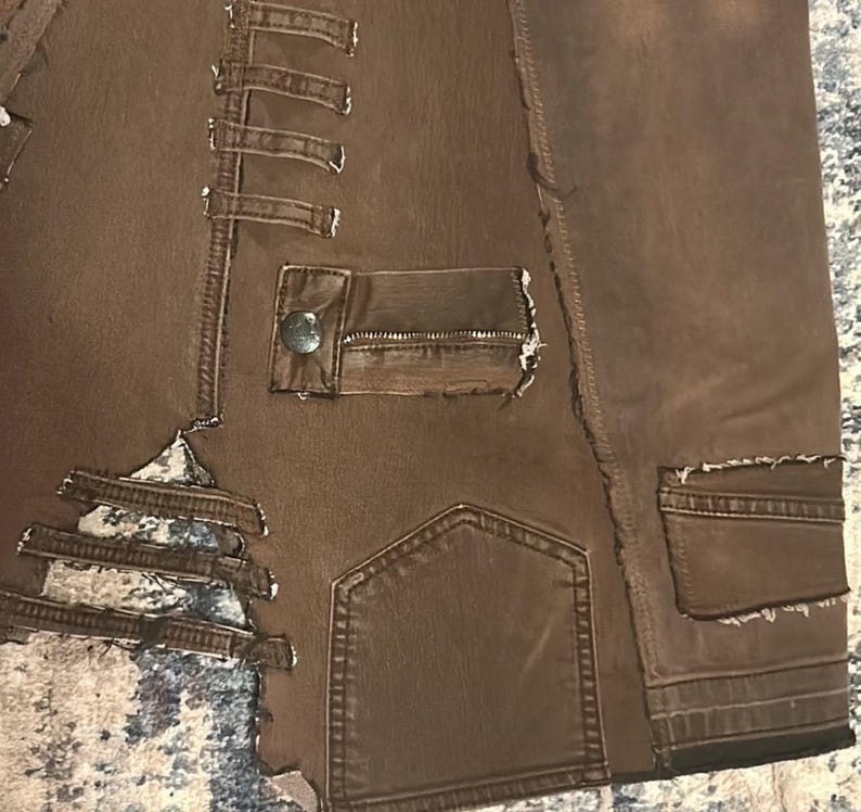 May include: Close-up of a brown jacket with multiple pockets and decorative stitching. The jacket has a distressed look with frayed edges and a button closure on one of the pockets. The fabric appears to be a sturdy material, possibly denim or a similar textile.