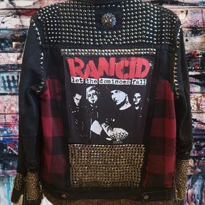 Puede incluir: Chaqueta vaquera negra con tachuelas plateadas, un panel a cuadros rojos y negros y un gráfico de la banda Rancid. La chaqueta presenta un acento de estampado de leopardo en la manga y un emblema decorativo en la parte superior de la espalda.