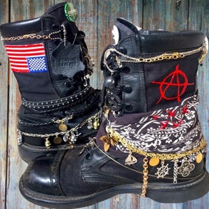 May include: Two black combat boots, adorned with chains, charms, and studs. One boot has an American flag patch, the other a red anarchy symbol. The boots are decorated with gold and silver-toned jewellery and a patterned fabric.