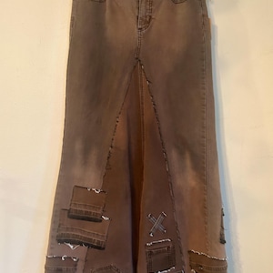 May include: Brown denim skirt with a high waist and a split front. The skirt features patchwork details and decorative stitching. The skirt is hanging on a wooden hanger against a neutral background.