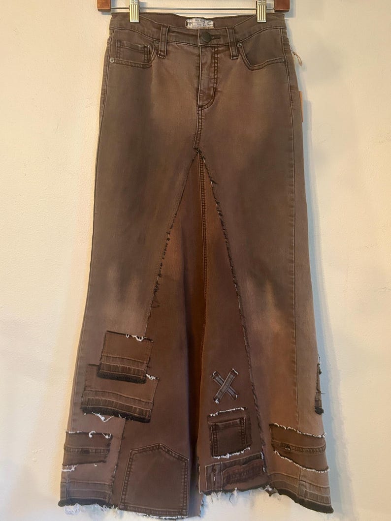 May include: Brown denim skirt with a high waist and a split front. The skirt features patchwork details and decorative stitching. The skirt is hanging on a wooden hanger against a neutral background.