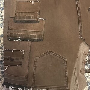 May include: Brown denim pants with multiple patches and distressed edges. The pants feature a back pocket and several sewn-on patches in varying sizes. The edges of the patches and the bottom hem are frayed.