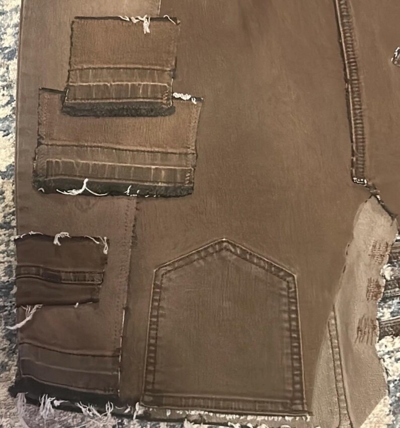 May include: Brown denim pants with multiple patches and distressed edges. The pants feature a back pocket and several sewn-on patches in varying sizes. The edges of the patches and the bottom hem are frayed.