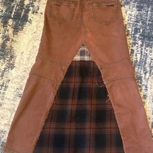 May include: Brown denim skirt with a plaid fabric panel. The skirt features a brown denim construction with back pockets and a plaid fabric insert in shades of black, brown, and tan. The skirt has a unique, repurposed design.