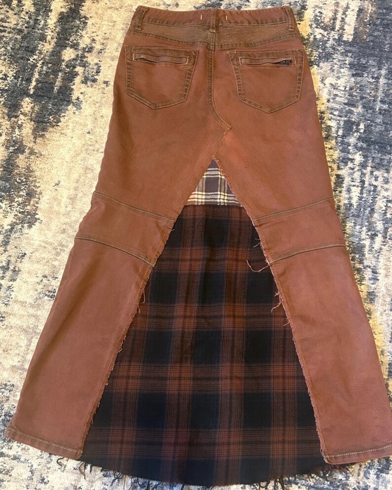 May include: Brown denim skirt with a plaid fabric panel. The skirt features a brown denim construction with back pockets and a plaid fabric insert in shades of black, brown, and tan. The skirt has a unique, repurposed design.