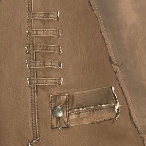 May include: Close-up of a brown leather jacket with decorative straps and a pocket. The jacket features a button closure on the pocket and visible stitching details. The leather has a distressed appearance.