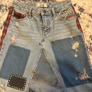 May include: Light blue denim skirt with patchwork design. Features distressed details, a red and brown plaid side stripe, and studded accents on the pocket. The skirt has a button closure and the number "24" on the waistband.