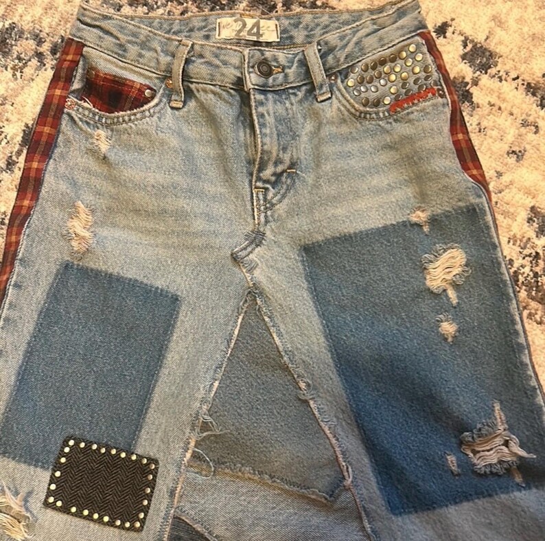 May include: Light blue denim skirt with patchwork design. Features distressed details, a red and brown plaid side stripe, and studded accents on the pocket. The skirt has a button closure and the number "24" on the waistband.