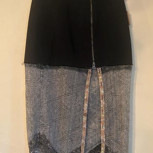 K&ouml;nnte beinhalten: Schwarzer Jeansrock mit silbernem Rei&szlig;verschluss und grauem Spitzen-Overlay. Der Rock hat einen Stoffstreifen mit Blumenmuster und Kn&ouml;pfen vorne sowie eine schwarze Spitzenborte am Saum.