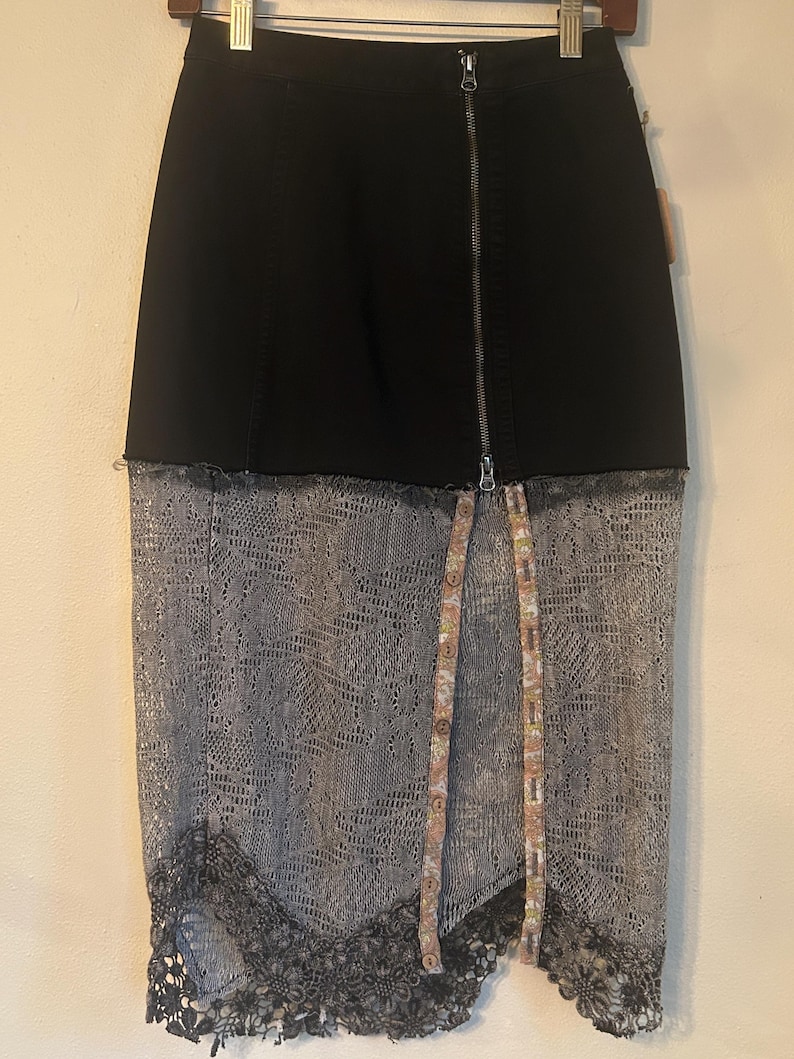 K&ouml;nnte beinhalten: Schwarzer Jeansrock mit silbernem Rei&szlig;verschluss und grauem Spitzen-Overlay. Der Rock hat einen Stoffstreifen mit Blumenmuster und Kn&ouml;pfen vorne sowie eine schwarze Spitzenborte am Saum.