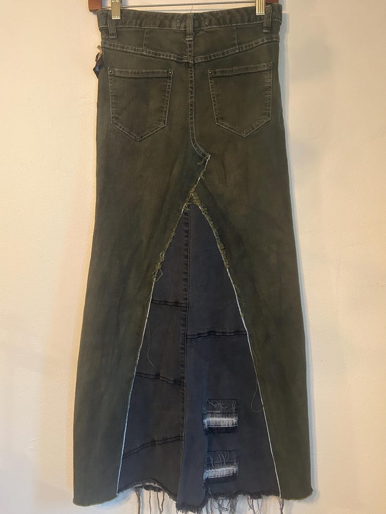 Puede incluir: Falda larga de mezclilla verde oscuro con cintura alta y dise&ntilde;o de espalda dividida. La falda presenta dos bolsillos traseros y un panel de mezclilla azul oscuro contrastante en la abertura, con parches desgastados y bordes deshilachados. La falda cuelga de una percha de madera.