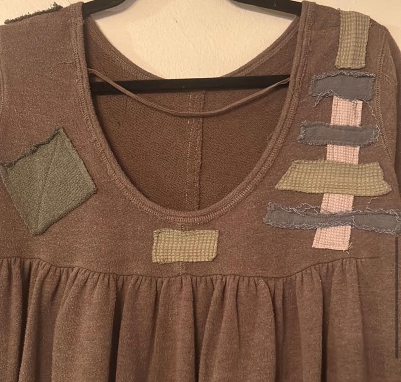 Może przedstawiać: Brązowy top z długim rękawem, okrągłym dekoltem i marszczoną talią. Top posiada ozdobne naszywki w odcieniach zieleni, szarości i r&oacute;żu. Naszywki są prostokątne i kwadratowe, dodając unikalny element designu do odzieży.