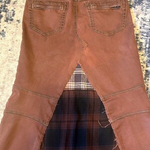 May include: A brown denim skirt with a patchwork design. The skirt features a brown denim front and back with pockets, and a plaid fabric insert at the bottom. The skirt has a unique, edgy style.