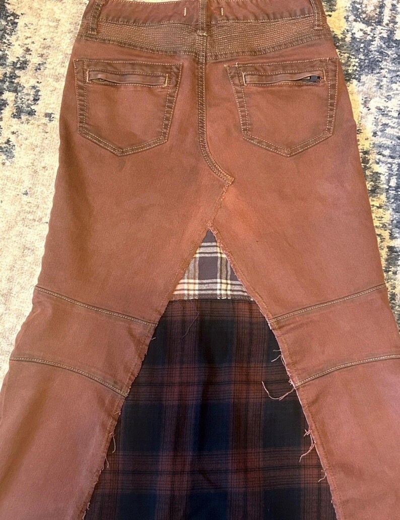 May include: A brown denim skirt with a patchwork design. The skirt features a brown denim front and back with pockets, and a plaid fabric insert at the bottom. The skirt has a unique, edgy style.