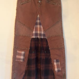 May include: A long, brown denim skirt with a patchwork design. The skirt features a plaid fabric panel in the center and at the bottom, with patches of plaid on the front. It has a button closure and zipper details.