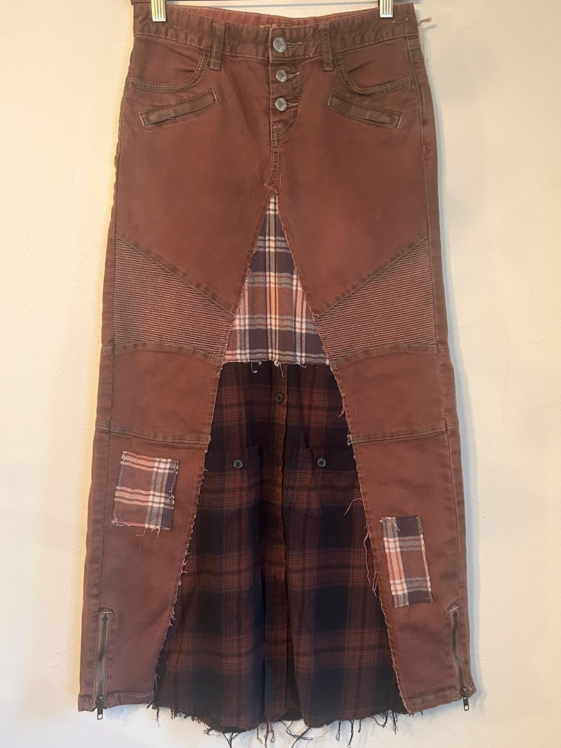 May include: A long, brown denim skirt with a patchwork design. The skirt features a plaid fabric panel in the center and at the bottom, with patches of plaid on the front. It has a button closure and zipper details.