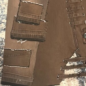 May include: Close-up of a brown leather jacket with multiple pockets and decorative stitching. The jacket has a distressed look with visible seams and a button closure. The pockets have a textured appearance.