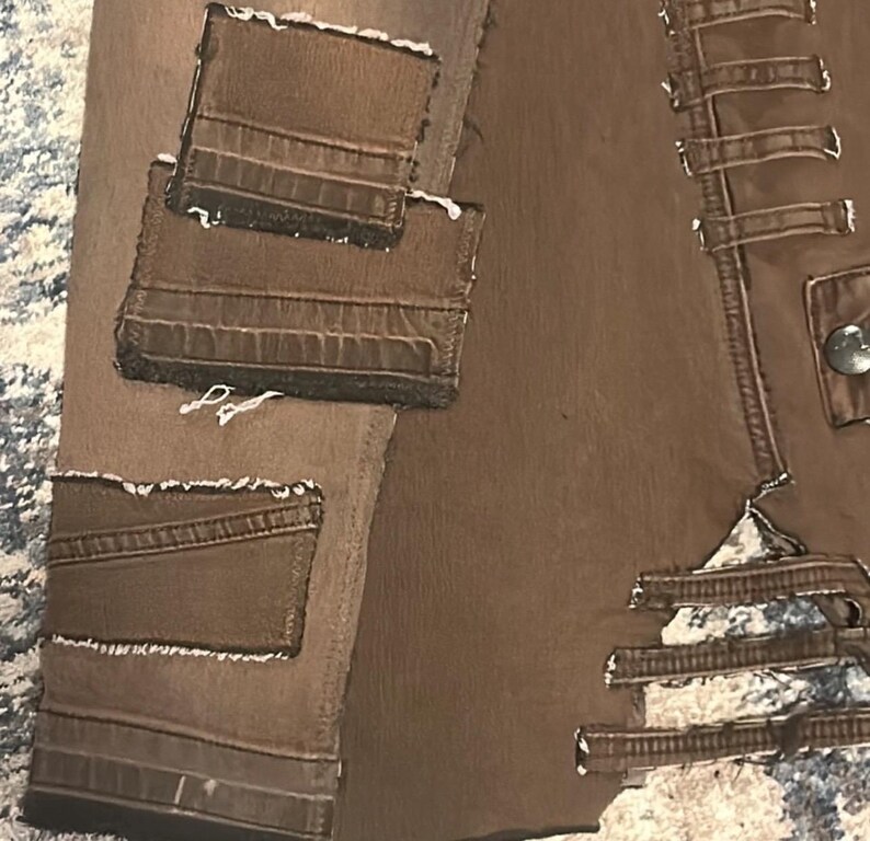 May include: Close-up of a brown leather jacket with multiple pockets and decorative stitching. The jacket has a distressed look with visible seams and a button closure. The pockets have a textured appearance.