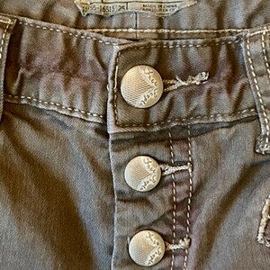 May include: Close-up of gray Free People jeans. The jeans have a button fly with three silver buttons. The brand label is visible, with the text "FREE PEOPLE" and "SIZE: W 25". White stitching accents the seams.
