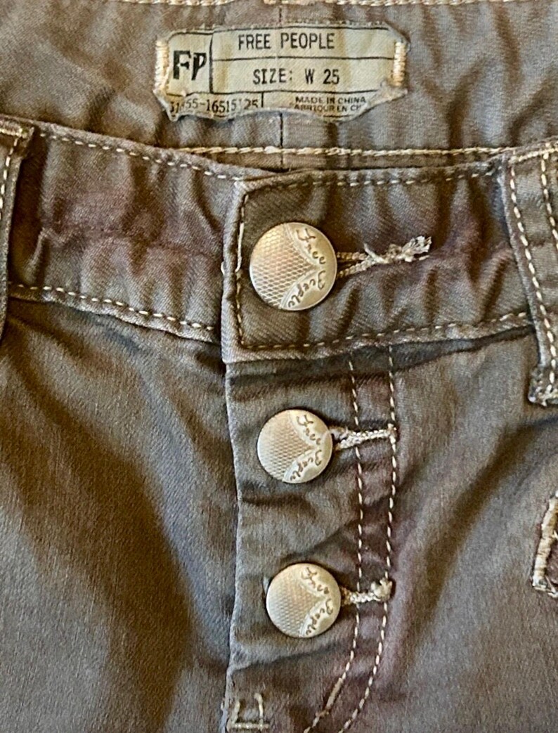 May include: Close-up of gray Free People jeans. The jeans have a button fly with three silver buttons. The brand label is visible, with the text "FREE PEOPLE" and "SIZE: W 25". White stitching accents the seams.