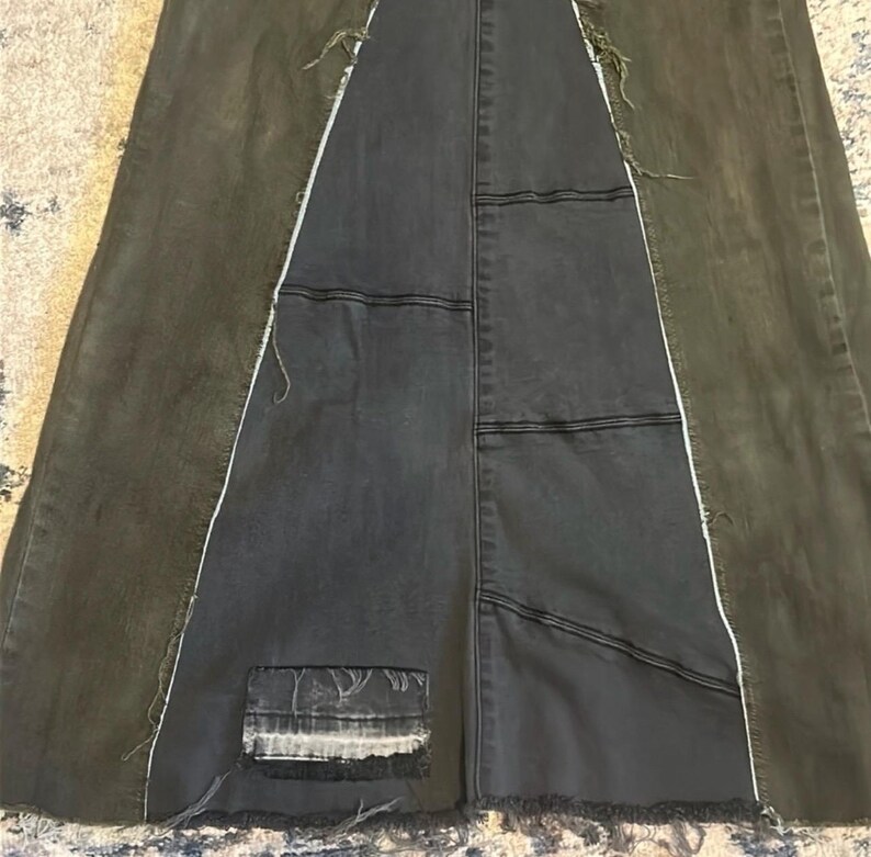 Puede incluir: Una falda larga de mezclilla oscura con un dise&ntilde;o de patchwork &uacute;nico. La falda presenta un panel central de mezclilla gris oscuro, flanqueado por paneles de mezclilla verde oliva. Las costuras blancas y los bordes deshilachados a&ntilde;aden detalles.