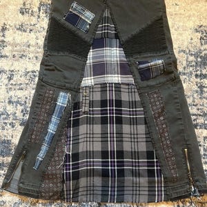 Puede incluir: Falda vaquera gris oscuro con dise&ntilde;o patchwork. La falda presenta un estampado de cuadros en tonos grises, negros y morados. Parches de tela azul y estampada est&aacute;n cosidos a la falda. La falda tiene cremalleras en los laterales y un estilo &uacute;nico y atrevido.