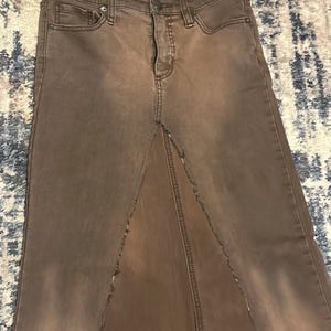 May include: A brown denim skirt with a high waist and a front slit. The skirt has a faded appearance and classic five-pocket styling. The skirt is made of a durable denim material.