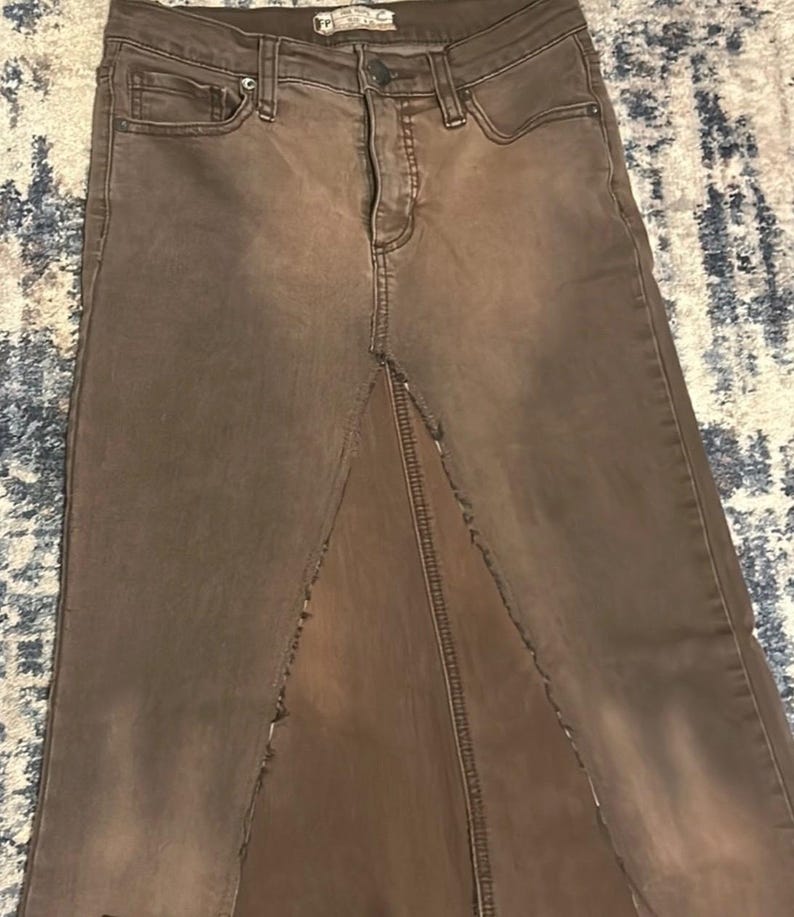 May include: A brown denim skirt with a high waist and a front slit. The skirt has a faded appearance and classic five-pocket styling. The skirt is made of a durable denim material.