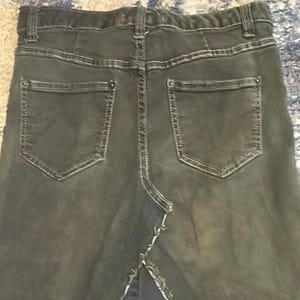 Puede incluir: Vista trasera de una falda vaquera gris oscuro con dos bolsillos traseros y costuras blancas. La falda tiene una abertura en el bajo y una cinturilla con trabillas. El tejido vaquero parece ligeramente descolorido.