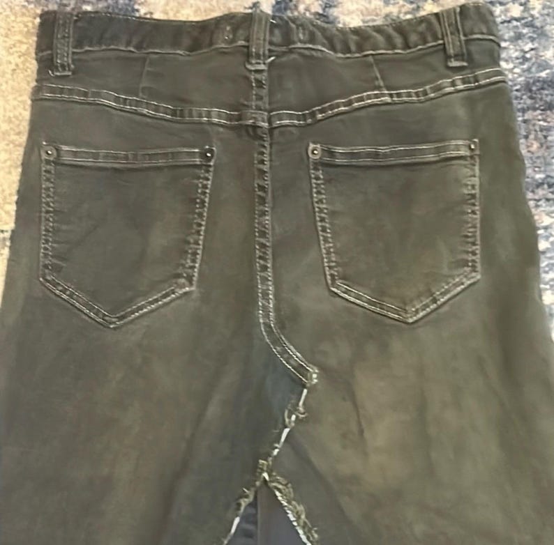 Puede incluir: Vista trasera de una falda vaquera gris oscuro con dos bolsillos traseros y costuras blancas. La falda tiene una abertura en el bajo y una cinturilla con trabillas. El tejido vaquero parece ligeramente descolorido.